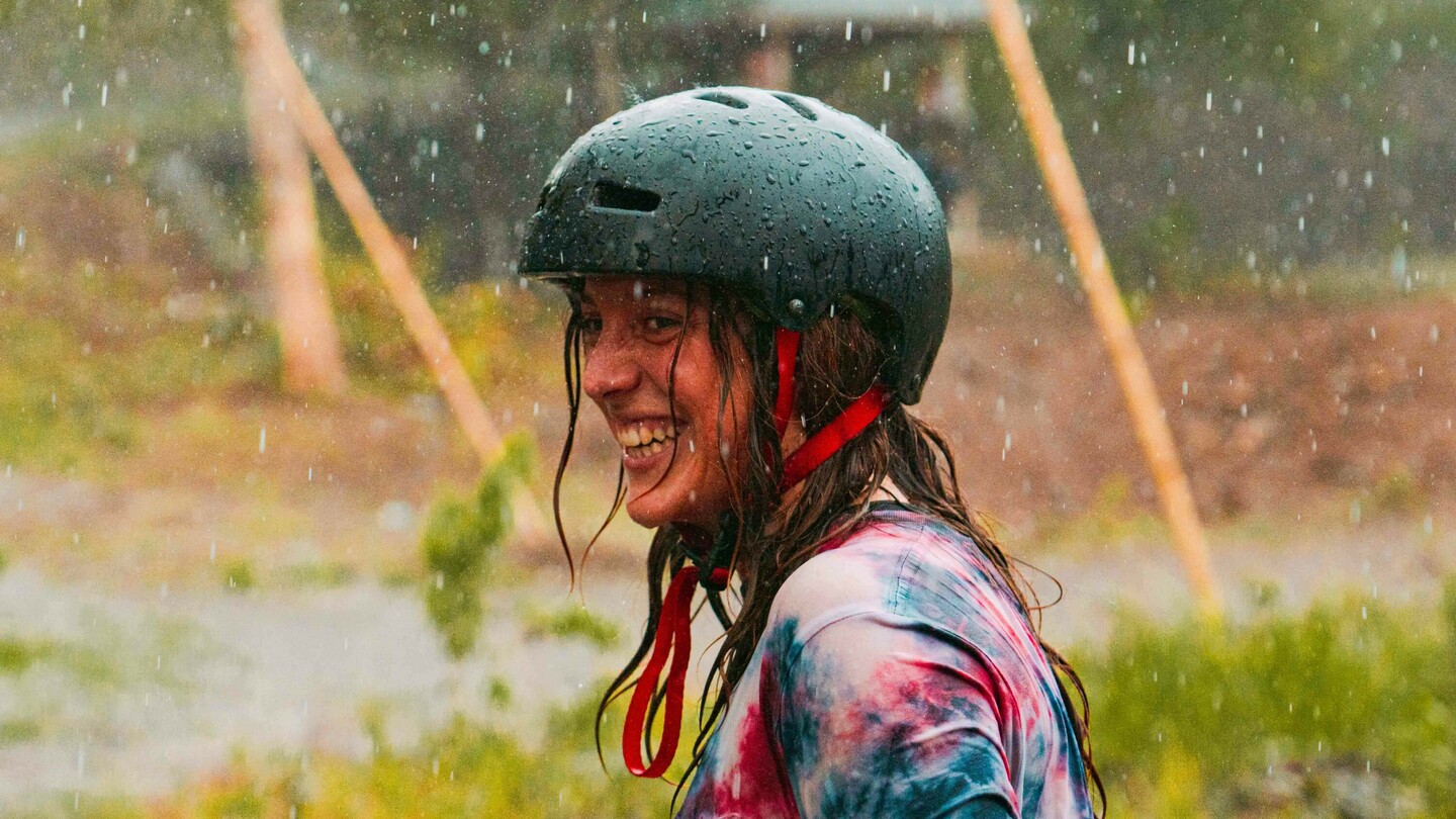 Anna Pixner, een downhill-skateboardster, staat lachend in de regen en draagt een zwarte helm en een kleurrijk tie-dye T-shirt. Op de achtergrond is een landelijke omgeving met een paviljoen en meerdere mensen te zien.