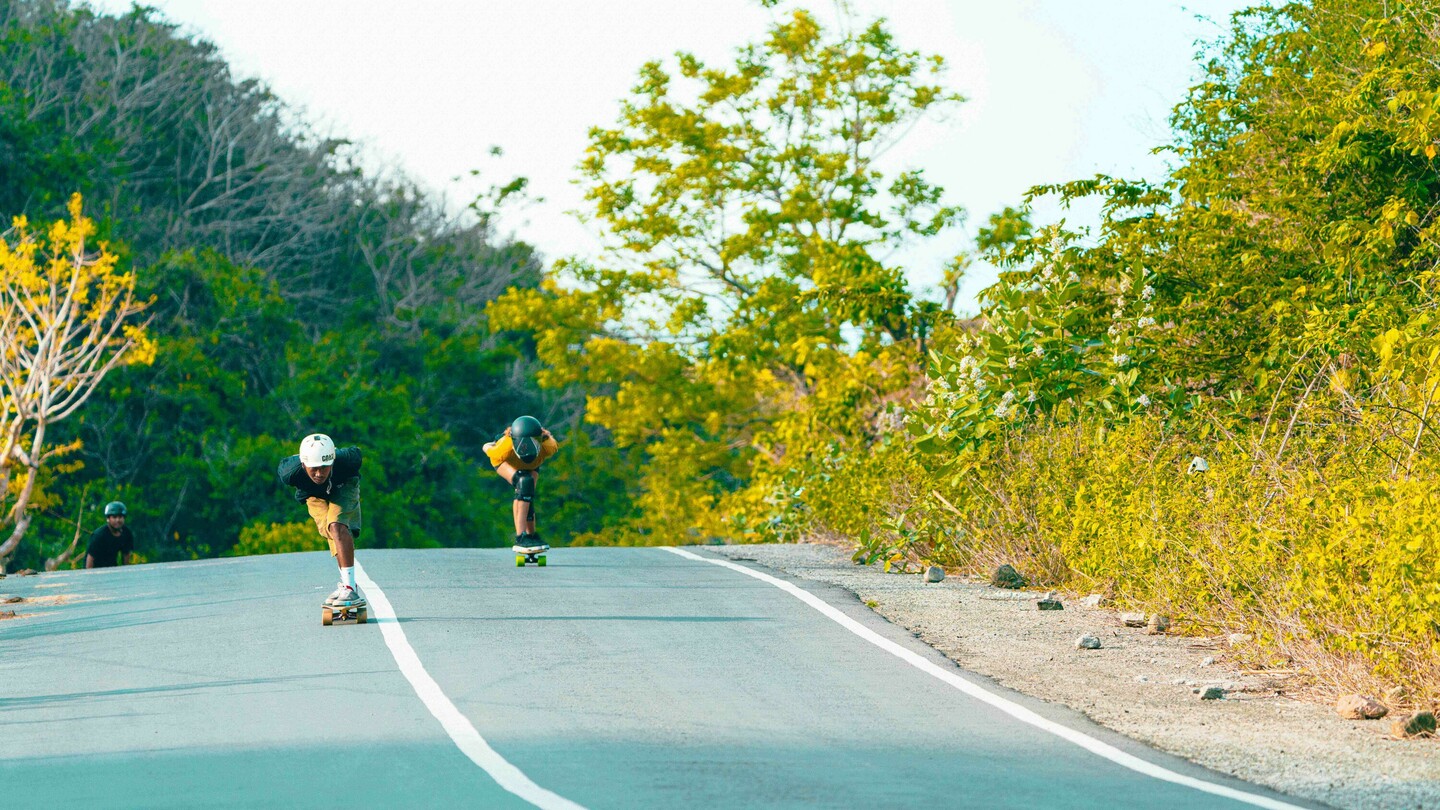 Meerdere downhill-skateboarders, waaronder Anna Pixner met een geel T-shirt en zwarte helm, rijden een landelijke, licht hellende weg af. De scène wordt omringd door weelderige vegetatie en bomen.