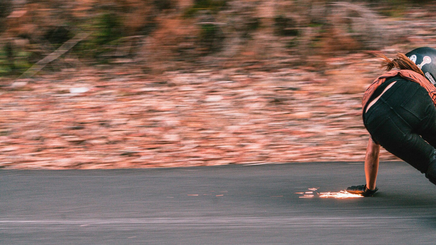 De Oostenrijkse downhill-skateboarder Anna Pixner is te zien terwijl ze met hoge snelheid op haar longboard rijdt. In een lage positie raakt ze met haar gehandschoende hand het asfalt, waardoor er vonken ontstaan. De bewegingsonscherpte op de achtergrond benadrukt de hoge snelheid en dynamiek van de scène, terwijl Anna een bocht op de weg maakt.