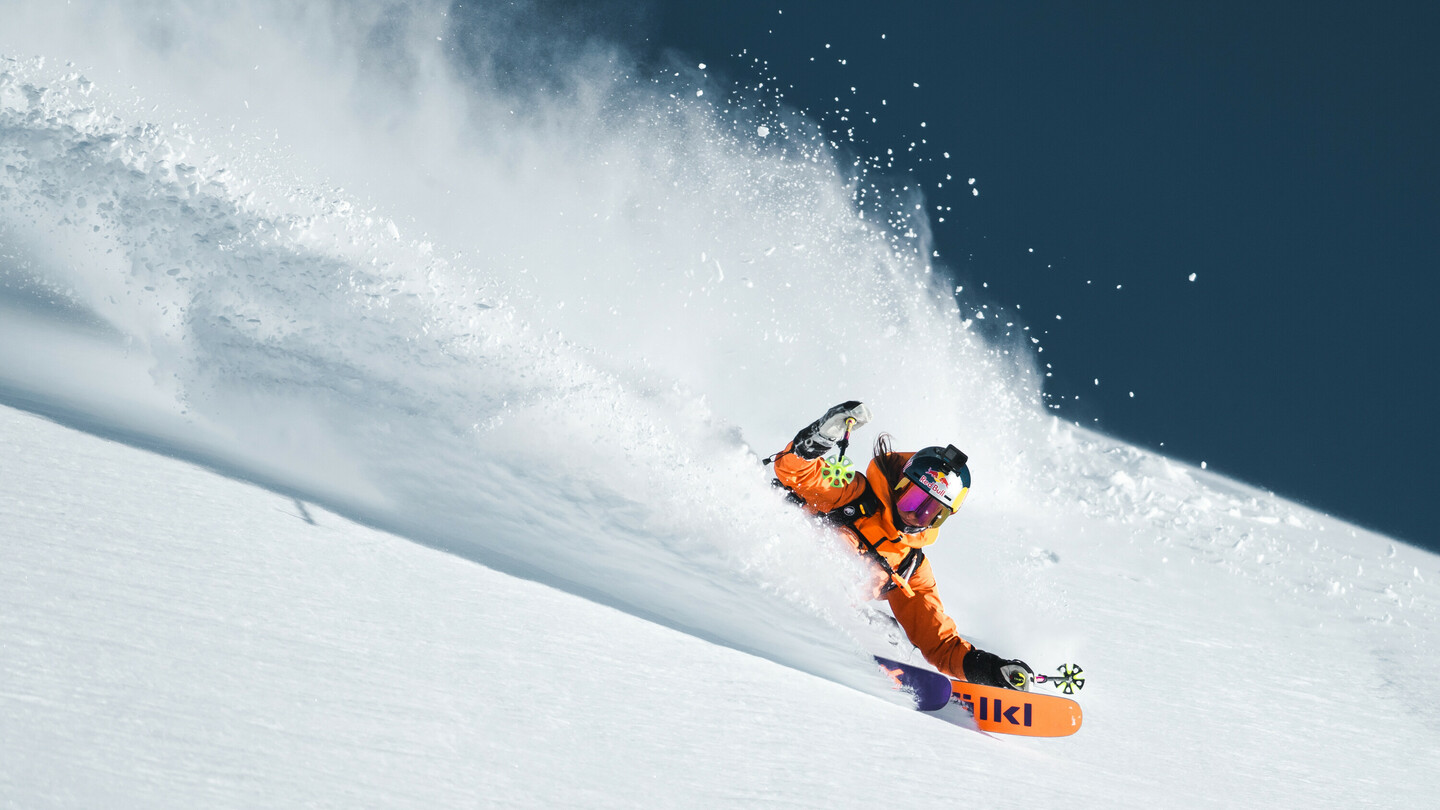 Vrouwelijke skiër in een oranje jas die dynamisch door diepe poedersneeuw skiet, met opspattende sneeuw op de achtergrond.