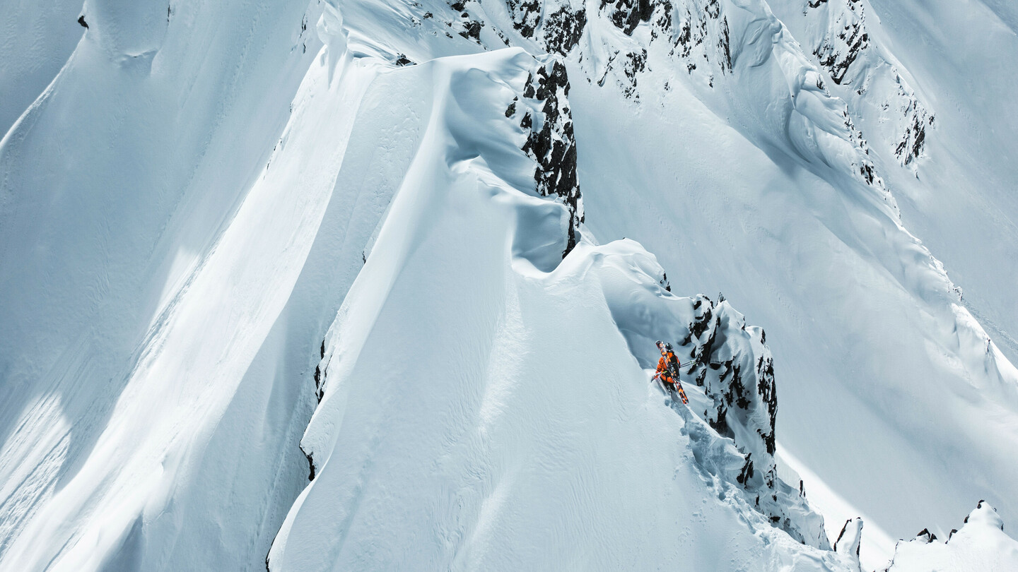 Skiër in een oranje jas navigeert voorzichtig langs een smalle, steile sneeuwrichel in een dramatisch berglandschap.Skiër in een oranje jas navigeert voorzichtig langs een smalle, steile sneeuwrichel in een dramatisch berglandschap.