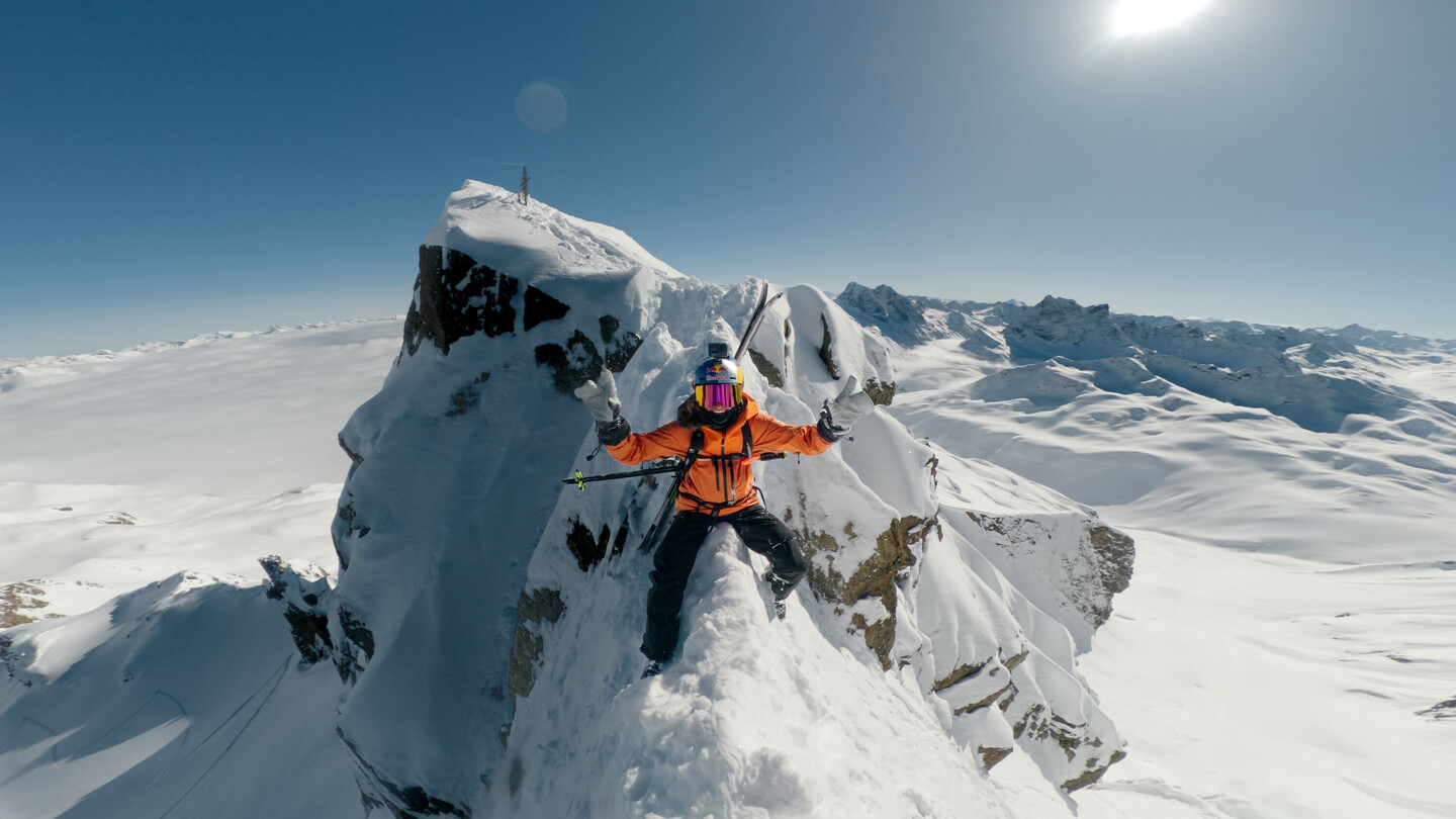 Skiër in een oranje jas balanceert op een smalle, met sneeuw bedekte rotsrichel, omringd door een spectaculair winterlandschap onder een heldere hemel.