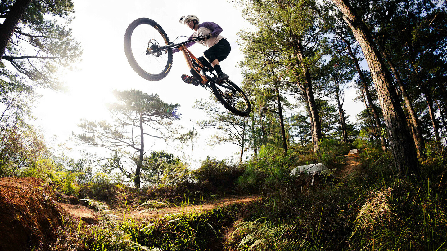 Een mountainbiker voert een spectaculaire sprong uit over een heuvel in een bosrijk gebied. De rijder draagt beschermende kleding en een helm, terwijl de zon door de bomen schijnt en het bos in warm licht baadt.