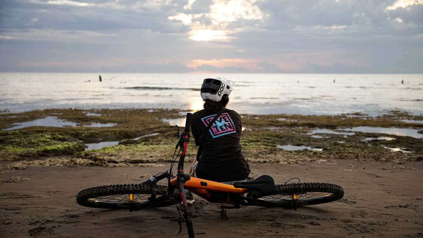 Een persoon met een helm zit op het strand naast een mountainbike en kijkt uit over de zee terwijl de zon achter de wolken ondergaat. De sfeer is rustig en ontspannen.