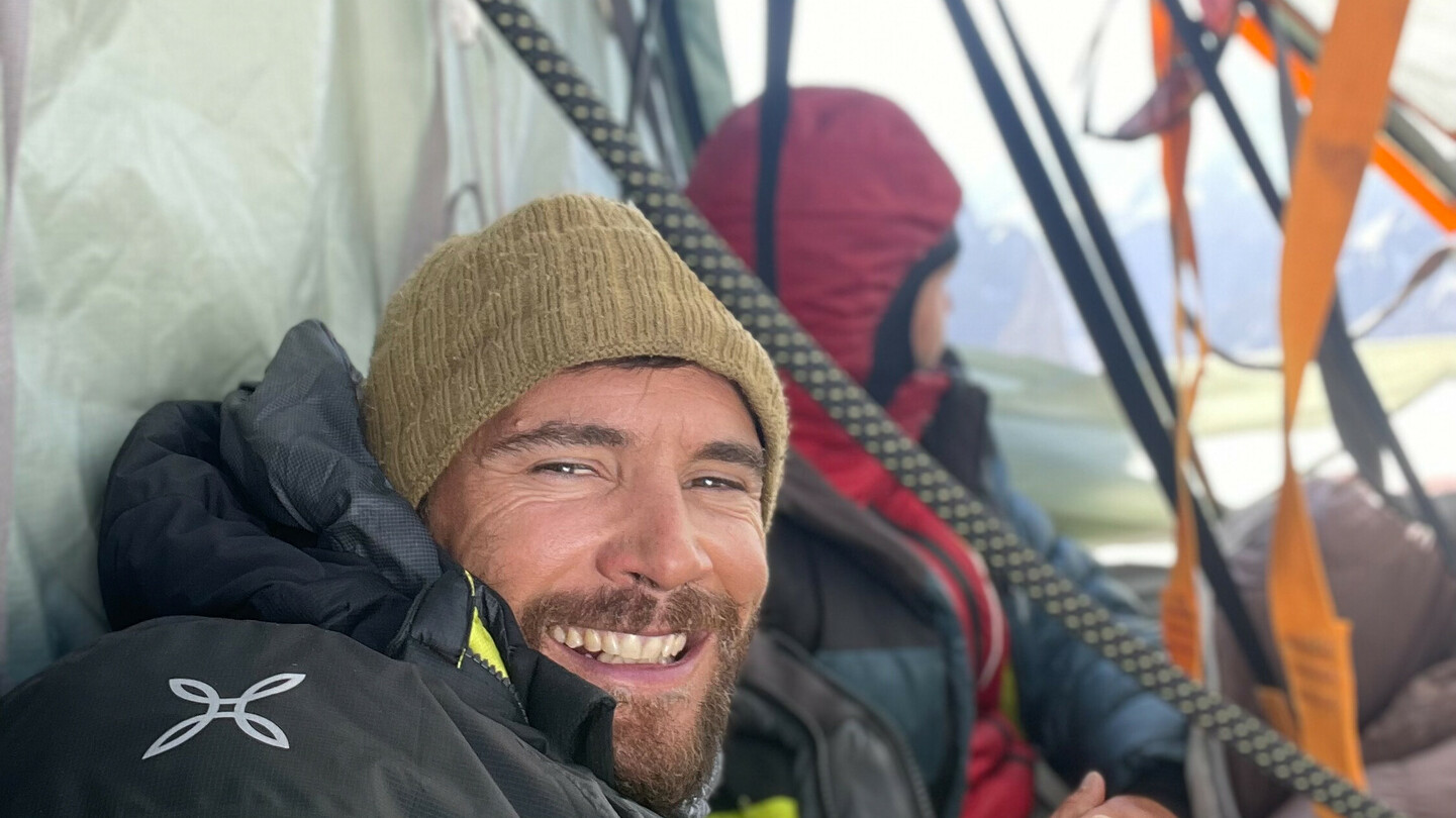 Eduard Marín glimlachend in een tent tijdens een bergexpeditie, warm ingepakt in outdoor kleding, met een andere klimmer op de achtergrond.