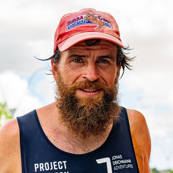 Portret van Jonas Deichmann, een man met een volle baard en een rode pet met het Bubba Gump Shrimp Co. logo. Hij draagt een donker tanktop.